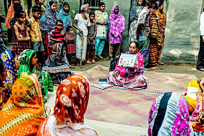 A female healthcare worker in Bangladesh crosslegged sits on a rug showing a with a circle of people a fact sheet about immunisation.