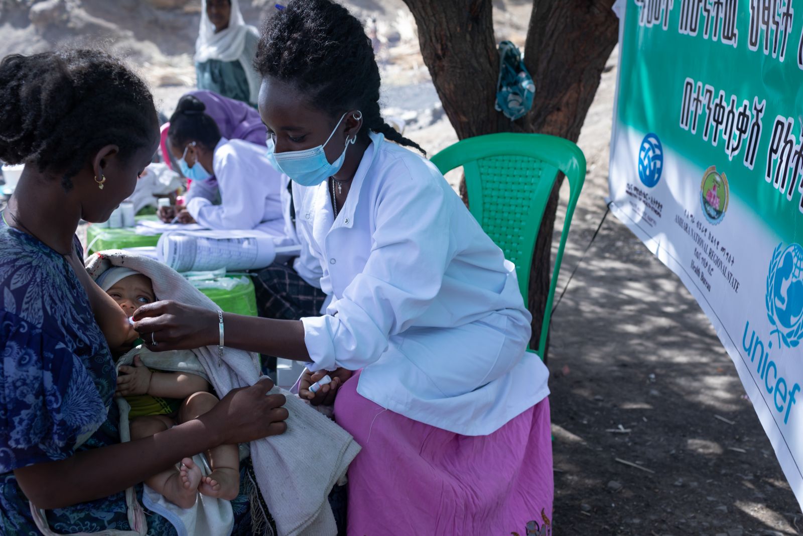 A black woman cradles a baby as another woman in a white medical coat gives the baby drops into its mouth